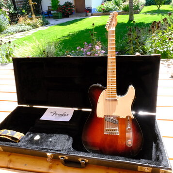 Elektrická kytara s tmeleým sunburst povrchem těla a světlým hmatníkem z javoru. Nástroj má bílý pickguard a na hlavě je viditelné logo Fender. Kytar…