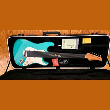 Tyrkysová elektrická kytara s bílým pickguardem v černém tvrdém kufru. Na hmatníku je nápis Xotic Guitars a na hlavě nástroje logo Xotic. V kufru se…