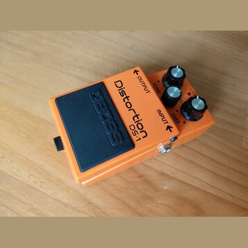 Oranžový efekt pedál s černým gumovým spínacím tlačítkem, na kterém je vyražený nápis BOSS. Na horní části je uveden název Distortion DS-1 a popisky…