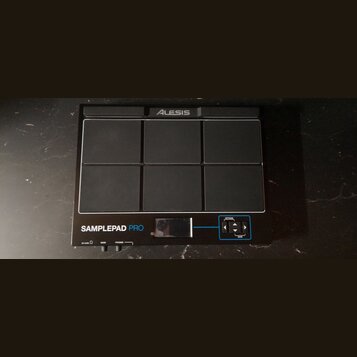 Černý elektronický sampling pad s šesti gumovými polštářky v provedení 2x3. V horní části je logo ALESIS a v dolní části nápis SAMPLEPAD PRO. V pravé…