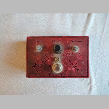 Červený kytarový efekt s nápisy Metal Simplex a Distortion. Na horní straně jsou tři ovládací prvky s popisky Gain, Tone a Volume. Povrch je výrazně…
