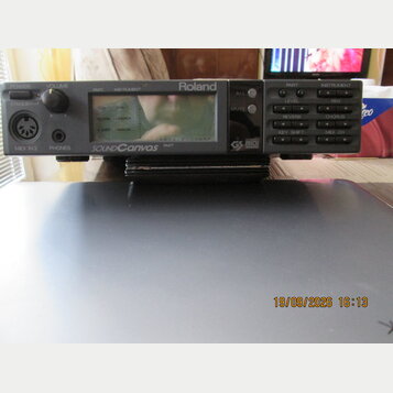 Šedý elektronický modul s displejem a ovládacími tlačítky. Na přední straně jsou nápisy Roland, SOUND CANVAS a GS MIDI. Jsou zde viditelné ovládací p…
