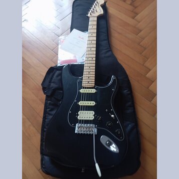 Černá elektrická kytara typu Stratocaster s bílým pickguardem a světlou hnedou hmatníkem. Na hlavě kytary je viditelné logo Fender a v pozadí leží pa…