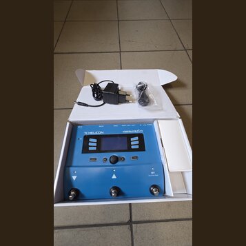 Modrý procesor vokálních efektů v bílé krabici. Na zařízení je nápis TC HELICON a model VoiceLive play. Součástí je napájecí adaptér a kabel v plasto…