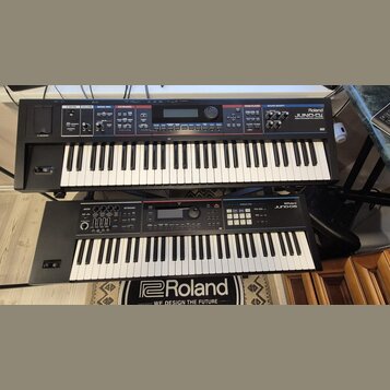 Dvě černé klávesové nástroje značky Roland umístěné nad sebou na stojanu. Horní instrument má nápis Roland JUNO-Di MOBILE SYNTHESIZER WITH SONG PLAYE…