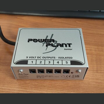 Stříbrný kovový napájecí adaptér s nápisem POWER PLANT Junior. Na horní straně je text 9 VOLT DC OUTPUTS - ISOLATED a čísla 1 až 5 nad odpovídajícími…