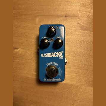 Modrý efekt pedál s nápisy FLASHBACK MINI a tc electronic. Na horní části jsou tři černé ovládací knoflíky s popisky DELAY, F. BACK a LEVEL. V dolní…