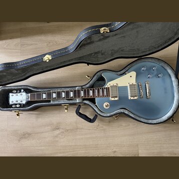 Modrá elektrická kytara v černém tvrdém kufru. Na hlavě je viditelné logo Epiphone. Kytara má dva humbuckery, bílý pickguard a čtyři ovládací knoflík…