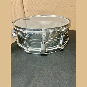 Stříbrný kovový snare drum s průhlednou blánou. Nástroj nemá žádné viditelné značky ani nápisy. Povrch kovu vykazuje drobné škrábance a známky opotře…
