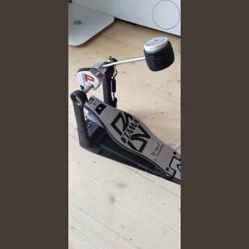 Krytý basový pedál s černým základem a stříbrným šlapadlem. Na šlapadle je viditelné logo TAMA a nápis The Legend in Innovation. Na horní části mecha…