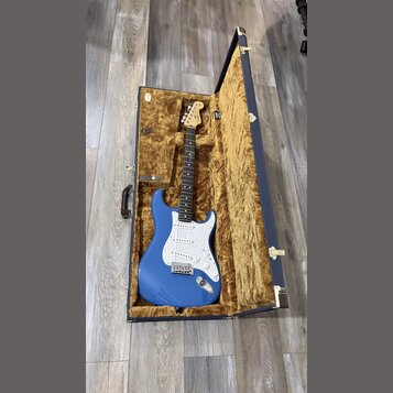 Modrá elektrická kytara typu Stratocaster s bílým pickguardem, uložená v otevřeném tmavomodrém kufru s hnědým plyšovým vnitřkem. Na hlavě kytary je v…