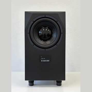 Černý studiový subwoofer v hranatém korpusu s matným povrchem. Na přední straně je umístěn velký kruhový reproduktor a pod ním ovládací panel s nápis…