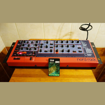 Červený rackový syntetizátor s nápisem nord rack VIRTUAL ANALOG. Na předním panelu jsou umístěny početné ovládací knofíky, tlačítka a displej s červe…