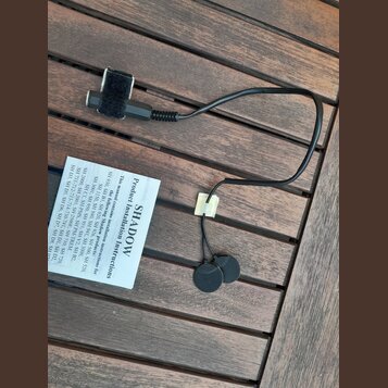 Černý kabel s dvěma kulatými lepicími senzory na koncích a suchým zipem v horní části. Vedle kabelu je bílý papírový návod s nápisem SHADOW Product I…