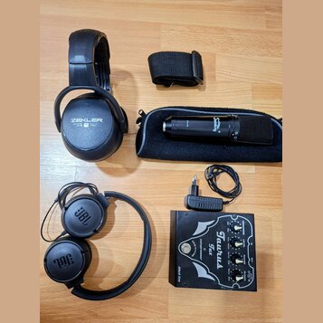 Na fotografii je sada audio vybavení: dvě černá sluchátka, mikrofon s pouzdrem a efektový pedál. První sluchátka mají nápis ZEKLER, druhá nesou logo…
