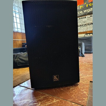 Černý reproduktor v hranatém korpusu s přední kovovou mřížkou s voštinovým vzorem. V dolní části mřížky je umístěno čtvercové logo s písmenem „K“ a n…