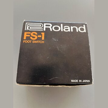Černá kartonová krabice s bílým logem Roland. Pod ním je oranžový nápis FS-1 a bílý text FOOT SWITCH. V pravém dolním rohu je bílý nápis MADE IN JAPA…