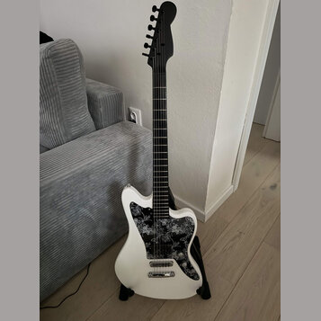 Bílá elektrická kytara s černým hmatníkem a černou hlavou. Kytara má černý pickguard s bílým květinovým vzorem a dva snímače typu single-coil. Na nás…
