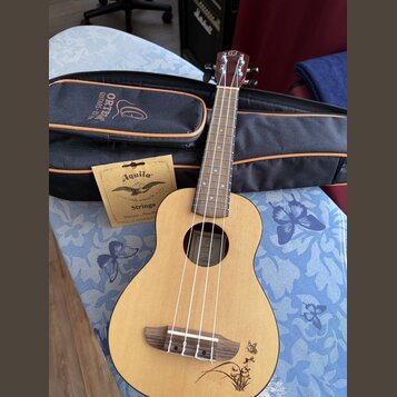 Světlý dřevěný ukulele s tmavým hmatníkem a čtyři struny. Na hlavě nástroje je viditelné logo ve tvaru stylizovaného písmene O. Součástí je černý pou…