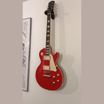 Červená elektrická kytara s tvarem single-cut, dvěma humbuckery a bílým pickguardem. Na hlavě hmatníku je viditelné logo "Kramer". Nástroj visí na ná…