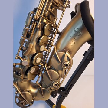 Detail saxofonu v matné zlaté barvě, který je umístěn na černém stojanu. Na korpu nástroje je vyryto logo SELMER PARIS a nápis MADE IN FRANCE. Pod ní…