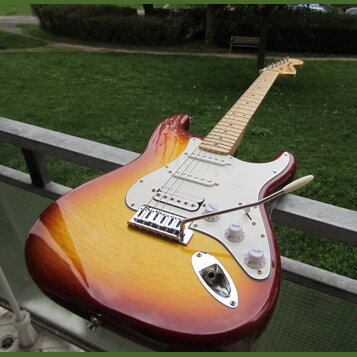 Elektrická kytara s tělem v barvě sunburst a bílým pickguardem. Na hlavě hředu je viditelné logo Fender. Nástroj má tři snímače, tři ovládací knoflík…