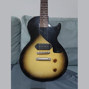 Elektrická kytara s tělem v žlutém gradientu (sunburst) a černým pickguardem. Nástroj má jeden humbucker v pozici mostu, dva zlaté ovládací knofíky a…
