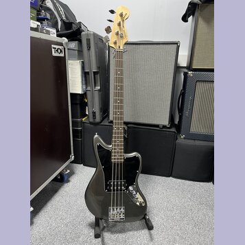 Tmavě šedá elektrická basová kytara s černým pickguardem a světle hnědým hmatníkem. Na hlavě nástroje je nápis JAGUAR BASS a logo Fender. Kytara stoj…
