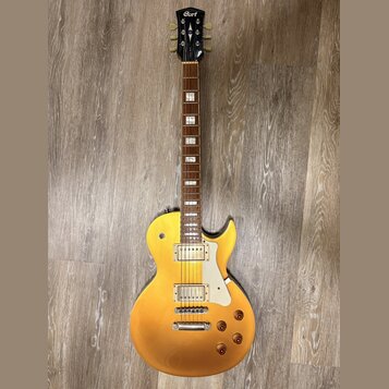 Zlatá elektrická kytara typu Les Paul s dvěma humbuckery a bílým plechtrátem. Na hlavě je vidět logo Cort. Kytara má čtyři ovládací knoflíky a hриф s…