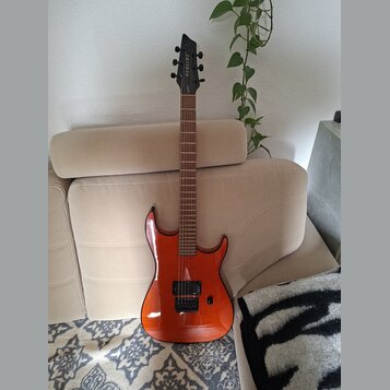 Elektrická kytara s oranžovým lakovaným tělem a hnědým hnedým hmatníkem. Na hlavě je vidět bílý nápis "Ibanez". Kytara má dva humbuckery, jeden ovlád…