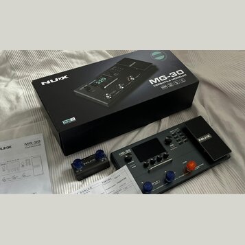 Na fotografii je kytarový procesor NUX MG-30 v černé barvě, doprovázený originální krabicí a dokumentací. Na přístroji je vidět nápis MG-30 VERSATILE…