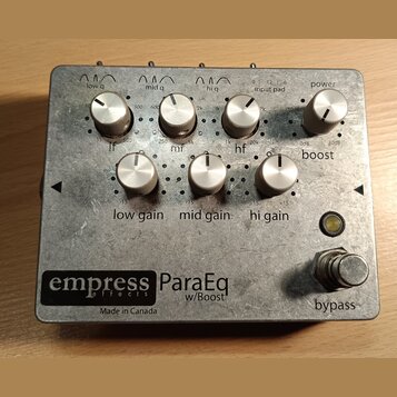 Kytarový efekt pedál v kovovém šasi stříbrné barvy. Na spodní části je černý štítek s nápisy „empress effects ParaEq w/Boost“ a pod ním text „Made in…