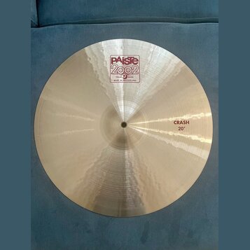 Kruhový činel z kovového materiálu s viditelným broušením. Na povrchu je červenobílé logo Paiste 2002 a nápisy TRADE MARK, MADE IN SWITZERLAND. V pra…