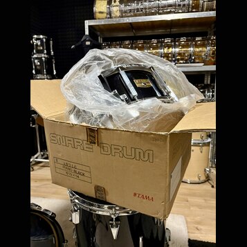 V kartonové krabici s nápisem SNARE DRUM a logem TAMA je umístěn černý malý buben zabalený v průhledné plastové fólii. Na korpusu nástroje je viditel…