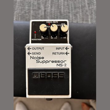 Kytarový pedál značky BOSS, model Noise Suppressor NS-2. Zařízení má bílé tělo s černým ovládacím panelem a černým spínacím pedálem. Na horní části j…
