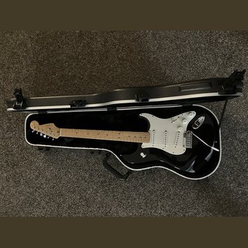 Černá elektrická kytara s bílým pickguardem v černém tvrdém kufru. Na hlavě kytary je viditelné logo Fender. Nástroj má světle hnědý javorový hmat a…