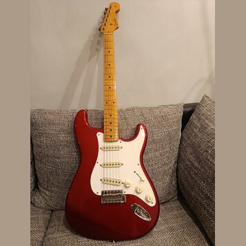 Elektrická kytara s červeným trupem, bílým pickguardem a hnedým javorovým hmatníkem. Na hlavě je viditelné logo Fender. Kytara má tři snímače typu si…