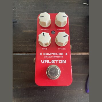 Červený kytarový efekt s bílými oválnými knoflíky. Na zařízení jsou nápisy OUTPUT, SUSTAIN, TONE a ATTACK, dále text COMPRINCE VINTAGE COMPRESSOR a z…