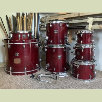 Sada červených bubnů s chromovaným kováním. Na korusech jsou viditelné loga „GRETSCH Custom Style“. Povrch největšího bubnu vykazuje v dolní části od…