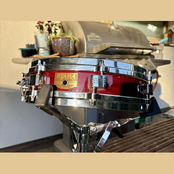 Červený snare drum s chromovanými obručmi a ladicími šrouby. Na korpusu je zlatý nápis TAMA a štítek s textem SNARE DRUM, LEGAL No. 200, MADE IN JAPA…