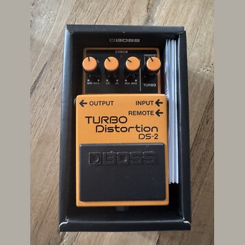 Kytarový efekt v oranžovém provedení s černým pedálem, umístěný v černé krabici značky BOSS. Na přístroji jsou nápisy TURBO Distortion DS-2, OUTPUT,…