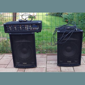 Dva černé reproduktory značky ALESIS s kovovými mřížkami. Na levém reproduktoru je umístěný černý mixážní pult s nápisy ALESIS a PAM. Na pultu jsou v…
