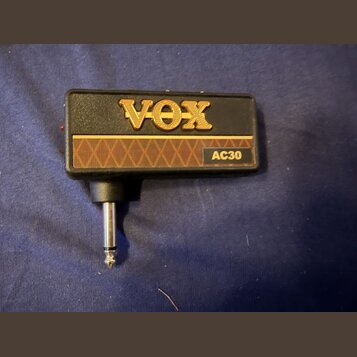 Černý obdélníkový adaptér s připojeným 6,3mm jack konektorem. Na horní straně je zlaté logo VOX a pod ním hnědý pruh s kosočtvercovým vzorem a nápise…