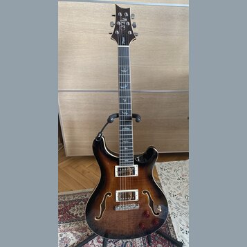 Polodublová elektrická kytara s tmavě hnědým gradientem (sunburst) a dvěma humbuckery. Na hlavě je bílý nápis "Cort" a menší text "Piezo". Hmatník ob…