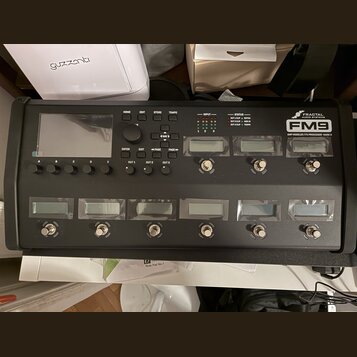 Černý kytarový procesor s displejem a ovládacími tlačítky. Na pravé straně je bílý nápis FRACTAL AUDIO SYSTEMS a model FM9 AMP MODELLER / FX PROCESSO…