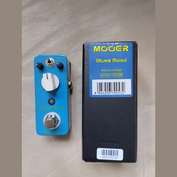 Modrý efekt pedál s bílým ovládacím knoflíkem, dvěma černými potenciometry a přepínačem. Na pedálu jsou nápisy „Blues Mood“ a „MOOER“. Vedle je černá…