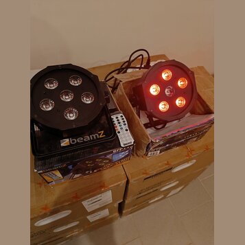 Dvě černé LED par lampy s šesti světelnými zdroji, z nichž jedna svítí červeně. Na základně levé lampy je bílý nápis beamZ. Pod přístroji jsou karton…