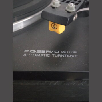 Detailní záběr na černý gramofon s nápisem F·G·SERVO MOTOR AUTOMATIC TURNTABLE. Na raménku je nasazen žlutý hloubkový systém s logem Audio-Technica.…