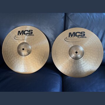Dvě mosazné činely s koncentrickým rýhováním, položené na modrém koženém povrchu. Oba mají černý nápis MCS a logo MEINL. Na levém činele je drobný ná…