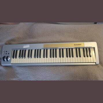 Stříbrný MIDI kontrolér s klaviaturní klávesou. Na horním panelu je zlatý pruh a nápis M-AUDIO KEYSTATION 61. V levé části jsou umístěny ovládací kno…